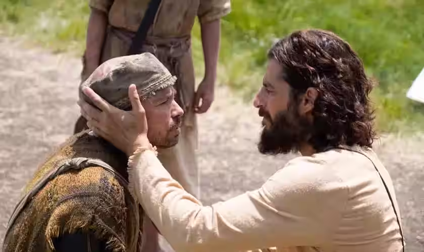 Jesus toca o rosto de um homem ajoelhado durante cena de cura na série The Chosen